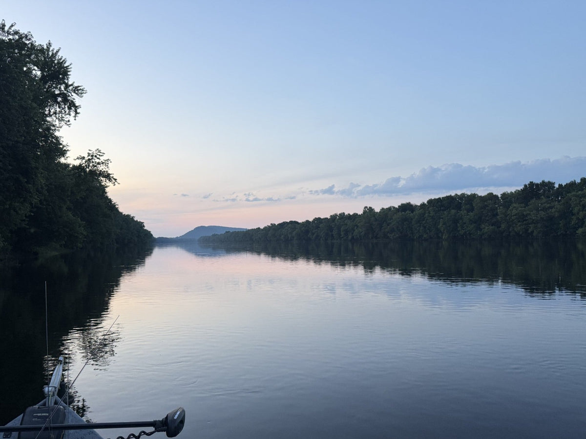The bow of a boat on a still, wide river under a pale blue and pink sky, with a wooded ridge in the distance