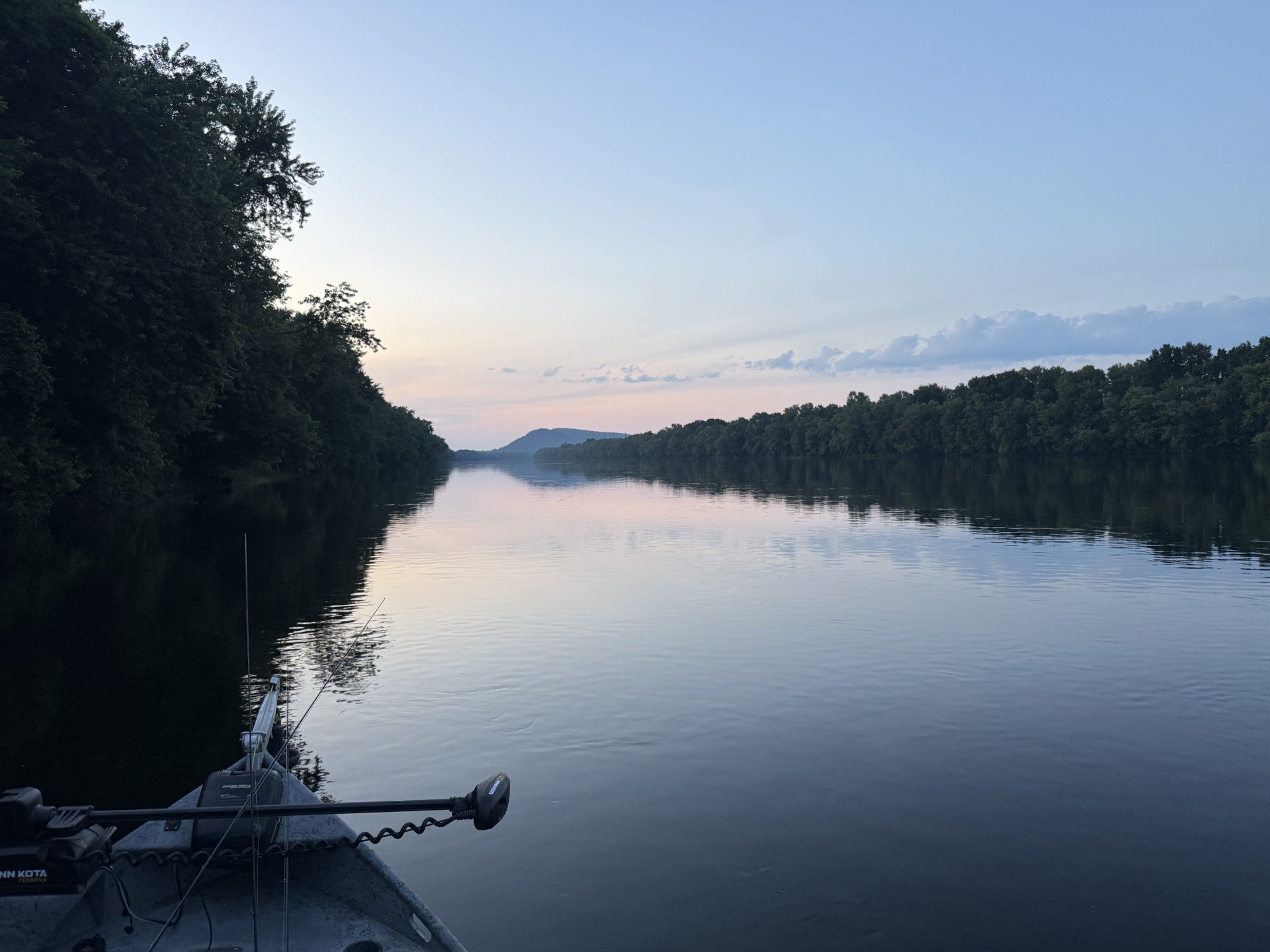Bow of a fishing boat on a calm river at dusk, tree-lined banks reflecting in still water, soft pink sky and a mountain ridge in the distance.
