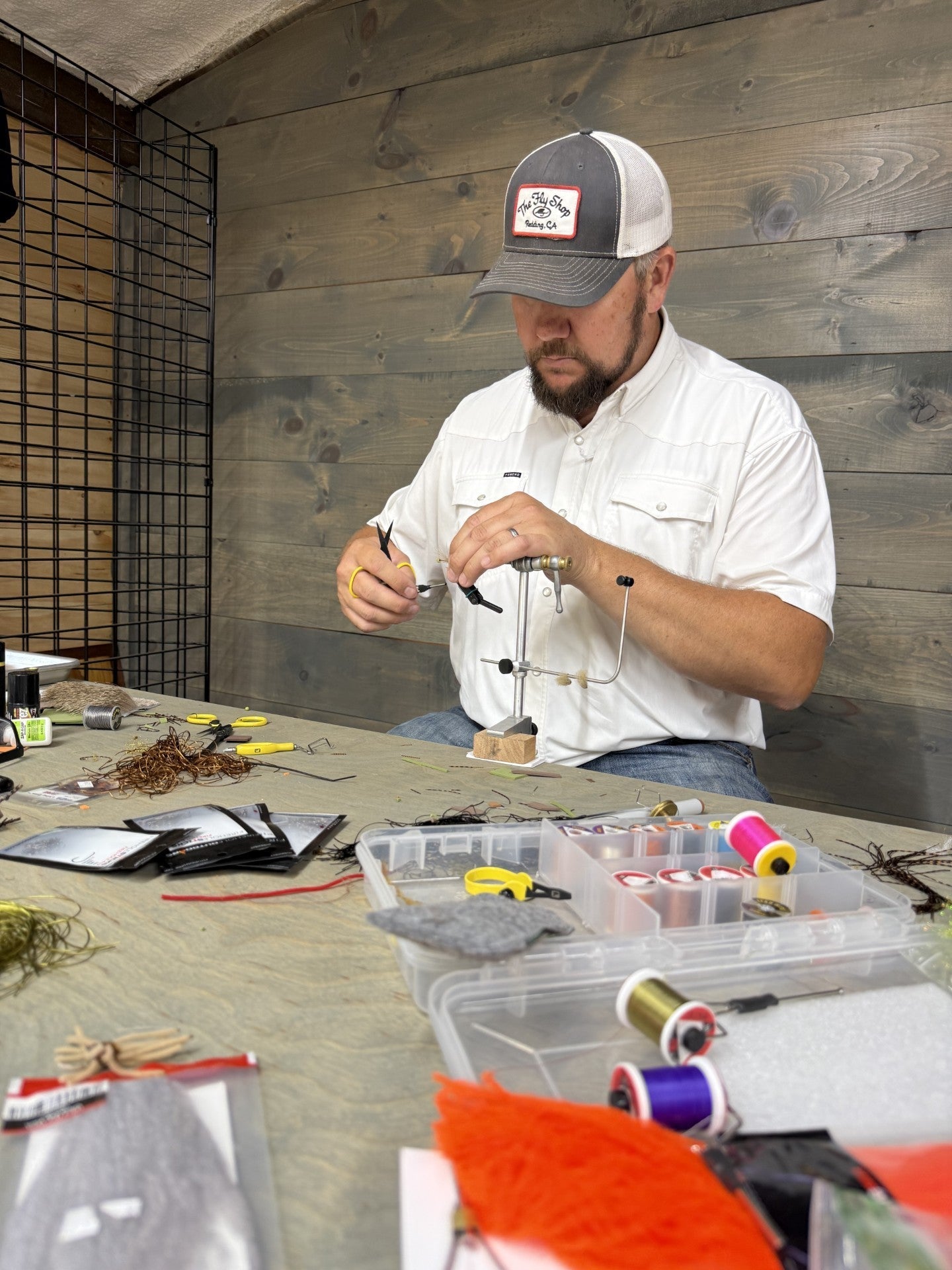 Fly tyer at the vise mid-tie, bench covered in thread spools, hackle, and materials