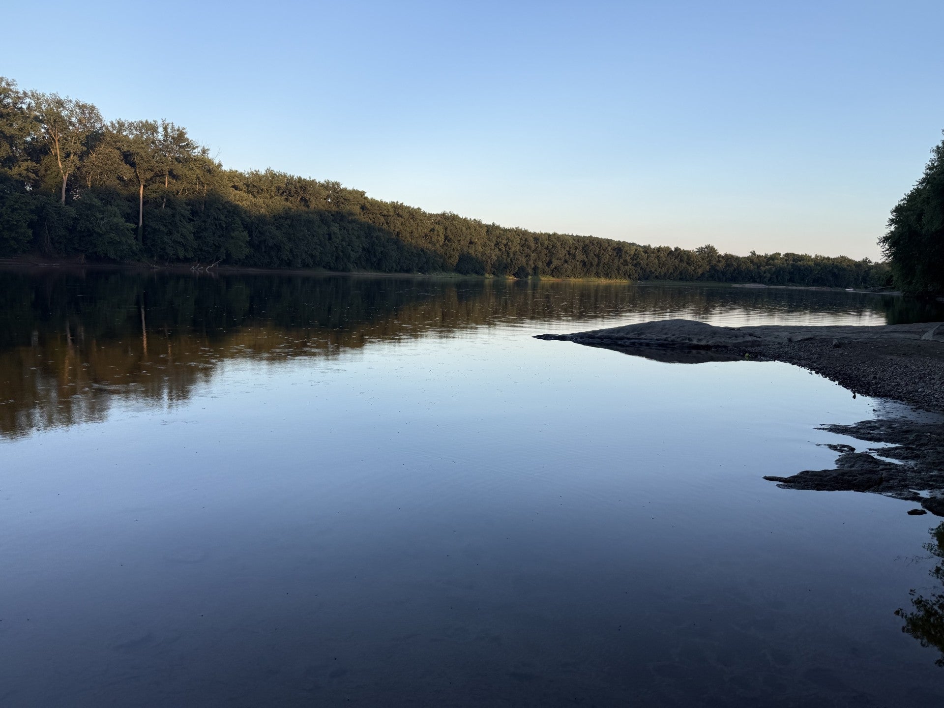 A calm river in low summer water at last light, trees along the far bank catching the evening sun, an exposed gravel bar in the foreground