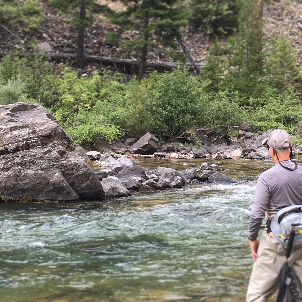 A fly angler with a sling pack and net stands at the edge of a riffle on a river in Montana, a large boulder across the current.