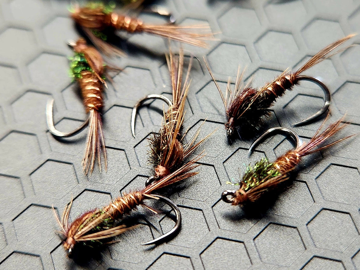 A scattering of pheasant tail nymphs with copper ribbing and green thoraxes on a grey tying mat